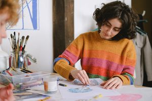 Concentrated female painter drawing sketches on paper with crayons while sitting at table near anonymous coworker in creative workshop during work