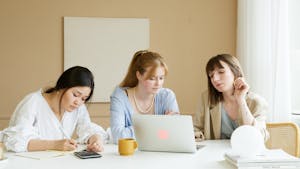 Three women working together in a modern office setting with a laptop and notes.