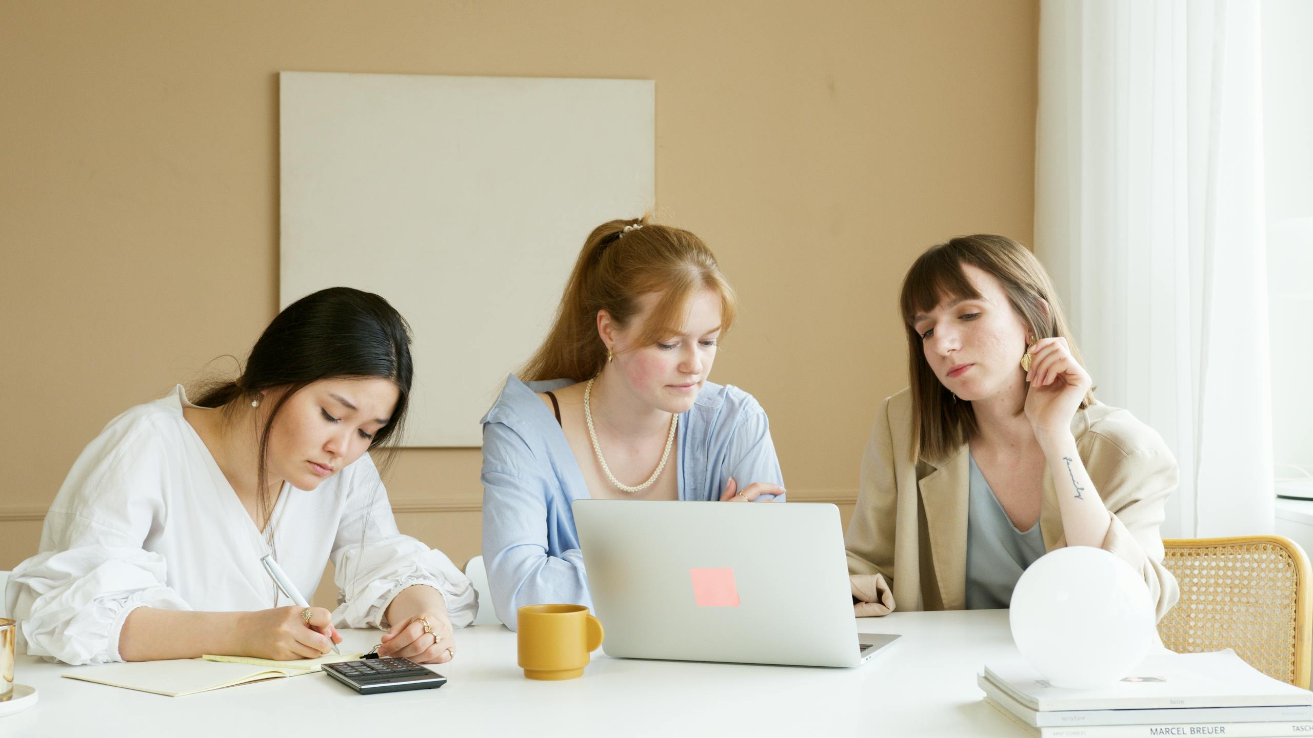 Three women working together in a modern office setting with a laptop and notes.